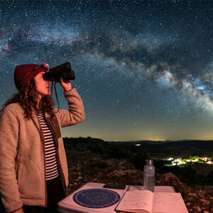 Una mujer observa el cielo nocturno y la Vía Láctea con prismáticos astronómicos junto a un planisferio iluminado con luz roja.