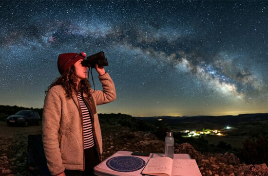 Una mujer observa el cielo nocturno y la Vía Láctea con prismáticos astronómicos junto a un planisferio iluminado con luz roja.