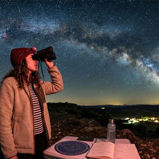 Una mujer observa el cielo nocturno y la Vía Láctea con prismáticos astronómicos junto a un planisferio iluminado con luz roja.